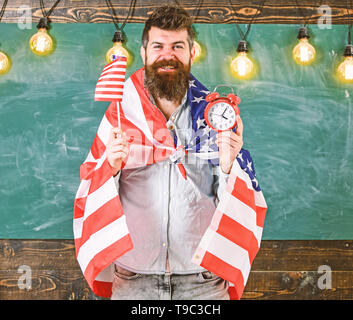 Mann mit Bart auf glückliches Gesicht hält Flagge der USA und die Uhr im Klassenzimmer, schwarzes Brett für den Hintergrund. Amerikanischen Bildungssystem Konzept. Amerikanische Lehrer mit amerikanischen Fahnen hält Wecker. Stockfoto