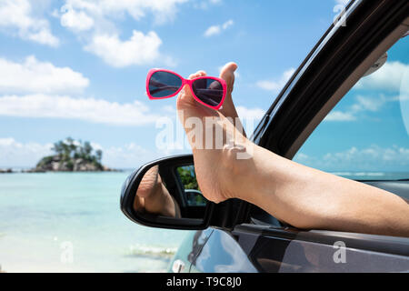 Close-up von Sonnenbrillen über die Füße der Frau aus dem Auto Fenster gegen den blauen Himmel Stockfoto