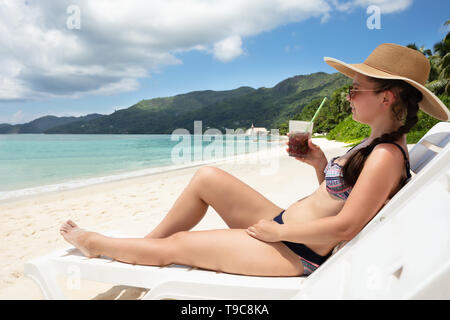 Junge Frau mit Hut sitzen auf weißen Liegestuhl genießen den Saft trinken am Strand. Stockfoto