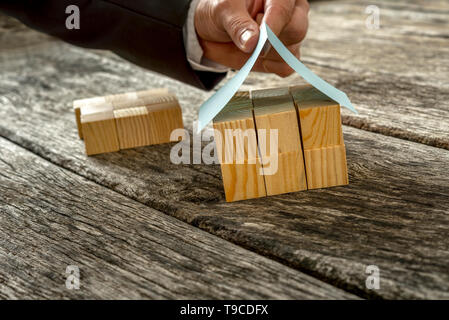 Nahaufnahme der männlichen Hand in eleganter Anzug, wenn Sie ein Papier-Dach auf Miniaturhaus aus Holz Würfelchen. Stockfoto