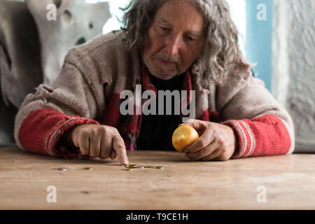 Senior Obdachloser in zerrissenen Pullover Essen alte Apple und Zählen von Euro Münzen auf einem alten Holztisch. Stockfoto