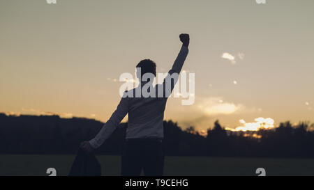 Ansicht von hinten von einem jungen Unternehmer stehen auf einer Wiese, wandte sich in Richtung auf ein Sonnenaufgang mit seinen Arm hoch in den Sieg. Stockfoto