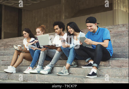 Gruppe von verschiedenen Jugendlichen mit Gadgets, sitzen auf der Treppe im Freien Stockfoto