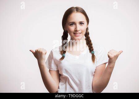 Schöne glückliche Schüler Mädchen lächelnd und mit Daumen nach oben Geste. Eine junge Frau trägt ein weisses T-Shirt und Pigtails, Geflecht und steht auf einem weißen zurück Stockfoto