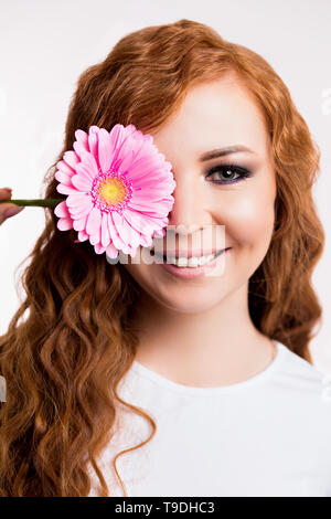 Nahaufnahme der schönen europäischen Frau mit roten Haaren, die Ihr Auge mit Rosa Gerbera Blume und lächelnd. Schönheit, Hautpflege, Frühling, urlaub Konzept Stockfoto