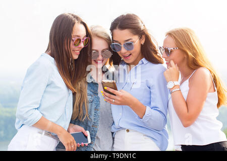 Vier schöne Frauen mit einem Smartphone. Die brünette Mädchen zeigt ihren Freunden ein Foto oder Video und jeder lacht, freut sich. Chat, das Internet, Technol Stockfoto