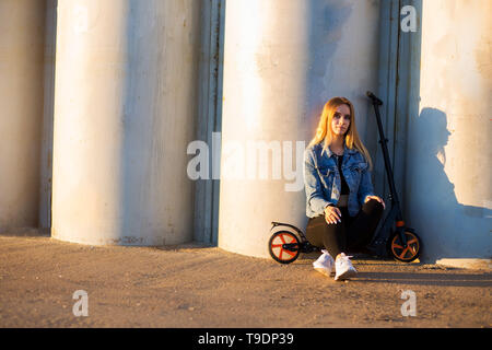 Sportliche Frau sitzt auf einem Roller in der Nähe der graue Wand, und wenn man die Kamera Stockfoto