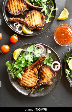 Gegrilltes Schweinefleisch Steaks mit Gemüse auf dem Teller über schwarzen Stein. Ansicht von oben, flach Stockfoto