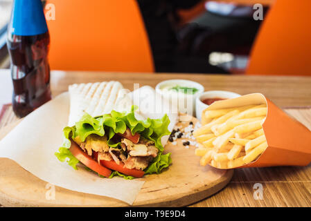 Shawarma und Pommes frites auf einer Holzplatte in einem Restaurant. Tortilla mit einem Drink in einem Cafe. Stockfoto