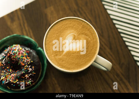 Kaffee mit einem Herz aus Milch und ein Donut auf einem Holztisch. Schlechtes Frühstück Stockfoto