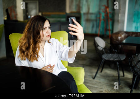 Eine junge, sympathische Frau, nicht eine dünne - vorangegangen Body Building, sitzt in einem gemütlichen Cafe, hält ein Smartphone in den Händen und hebt sich. Stockfoto