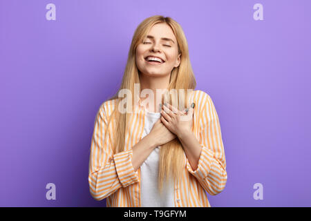 Art freundlich super Lachende junge Frau mit geschlossenen Augen hält beide Handflächen auf Brust, Dankbarkeit und gutes Gefühl über blauen Hintergrund isoliert. Studio gedreht. Stockfoto