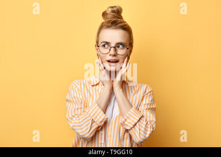 Stilvolle positive blonde Mädchen in der Runde Gläser mit hairbun in legere Kleidung, mit Palmen an den Backen, über Gelb Hintergrund isoliert. Verkauf, schockiert Preise Stockfoto