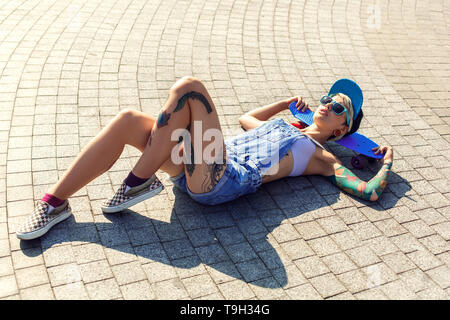 Junge alternative Mädchen skater tragen Cap und Sonnenbrille liegen auf der Straße Kopf auf Penny board entspannt Top View Stockfoto