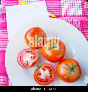 Frische reife rote Tomaten Tomaten in Scheiben geschnitten und mit Wassertropfen und einem grünen Stiel auf eine weiße Platte, neben einem Blatt Notepad schreiben und Steuerung Stockfoto