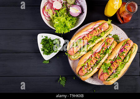 Hot Dog mit Wurst, Gurken, Rettich und Salat auf dunklem Hintergrund. Sommer hotdog. Ansicht von oben Stockfoto