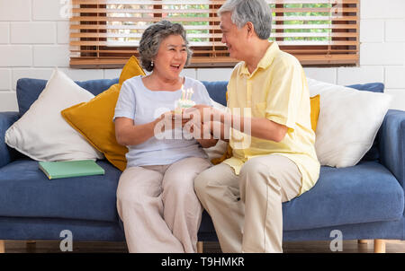 Asiatische älterer Mann überraschen ältere Frau mit Geburtstag Kuchen im Wohnzimmer zu Hause. Altern zu Hause Konzept. Stockfoto