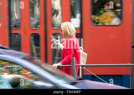 Belgrad, Serbien - April 22, 2019: junge blonde Frau an Ihr Mobiltelefon, während an der Bushaltestelle auf der Straße warten an einem sonnigen Frühlingstag mit Stockfoto