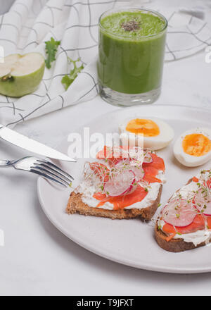 Toast mit Frischkäse und Lachs, hartgekochte Eier und grünen Smoothie. Gesundes Frühstück auf dem weißen Tisch Stockfoto
