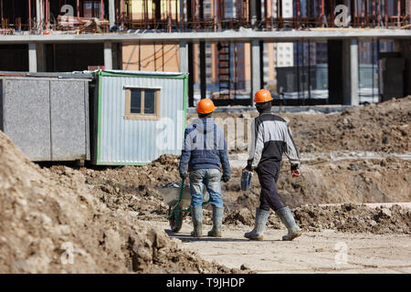 Zwei Arbeiter in Helme sind eine Schubkarre auf dem Hintergrund eines Gebäudes Schuppen angetrieben. Stockfoto