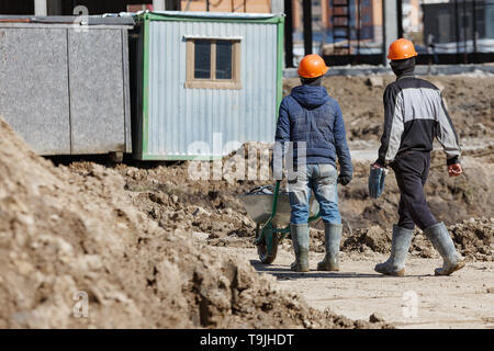 Zwei Arbeiter in Helme sind eine Schubkarre auf dem Hintergrund eines Gebäudes Schuppen angetrieben. Stockfoto