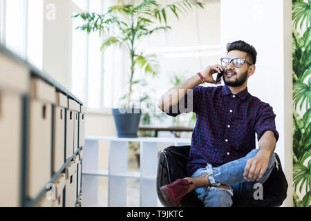 Lächelnd indischer Geschäftsmann sitzen auf einem Bürostuhl und Berufung auf dem Smartphone in modernen Büro Stockfoto