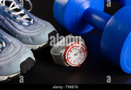 Ein Paar Turnschuhe, zwei blaue Kurzhanteln und eine moderne Uhr auf einem dunklen Hintergrund Stockfoto