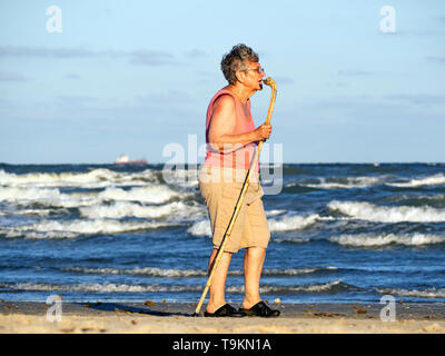 Eine ältere kaukasische Frau mit einem Stock Spaziergänge neben dem Golf von Mexiko am Strand während der Goldenen Stunde in Port Aransas, Texas USA. Stockfoto