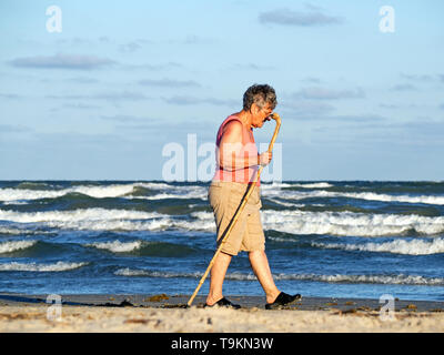 Eine ältere kaukasische Frau mit einem Stock Spaziergänge neben dem Golf von Mexiko am Strand während der Goldenen Stunde in Port Aransas, Texas USA. Stockfoto