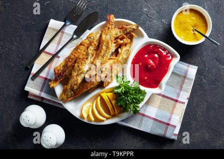 Gebratene kleine capelins mit Tomatensauce, in Scheiben geschnittenen Zitrone auf einer Platte auf einen konkreten Tisch mit Messer und Gabel, Ansicht von oben, Stockfoto