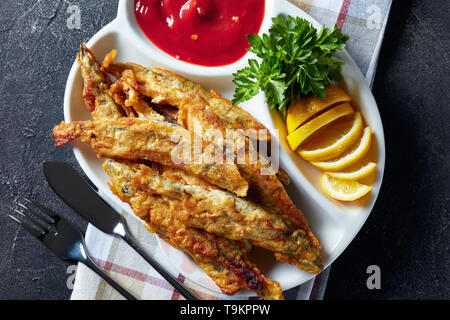 Gebratene kleine capelins mit Tomatensauce, in Scheiben geschnittenen Zitrone auf einer Platte auf einen konkreten Tisch mit Messer und Gabel, Ansicht von oben, flatlay, close-up Stockfoto