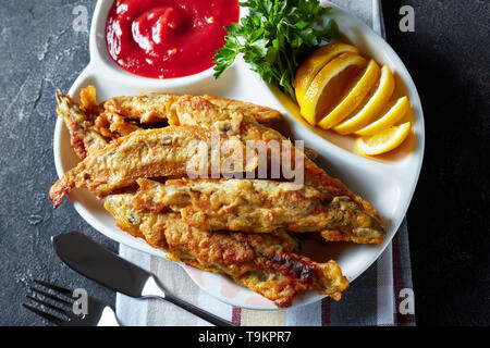 Gebratene kleine capelins mit Tomatensauce, in Scheiben geschnittenen Zitrone auf einer Platte auf einen konkreten Tisch mit Messer und Gabel, Ansicht von oben, flatlay, close-up Stockfoto