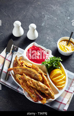 Gebratene kleine capelins mit Tomatensauce, in Scheiben geschnittenen Zitrone auf einer Platte auf einen konkreten Tisch mit Messer und Gabel, vertikale Ansicht von oben, leeren Raum Stockfoto