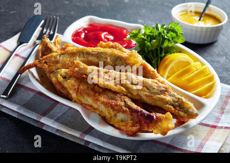 Gebratene kleine capelins mit Tomatensauce, Zitrone auf einem Schild auf einen konkreten Tisch mit Messer und Gabel in Stücke oder Scheiben geschnitten, horizontale Ansicht von oben, close-up Stockfoto