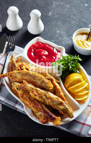 Gebratene kleine capelins mit Tomatensauce, Zitrone auf einem Schild auf einen konkreten Tisch mit Messer und Gabel in Stücke oder Scheiben geschnitten, vertikale Ansicht von oben, close-up Stockfoto