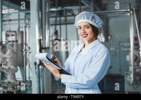 Eine junge schöne Mädchen in weißen Overalls macht sich Notizen in einer Tablette auf dem Hintergrund der Ausrüstung eines Nahrungsmittel- Anlage. Die Qualitätskontrolle in der Produkt Stockfoto