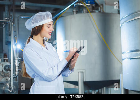 Eine junge schöne Mädchen in weißen Overalls macht sich Notizen in einer Tablette auf dem Hintergrund der Ausrüstung eines Nahrungsmittel- Anlage. Die Qualitätskontrolle in der Produkt Stockfoto