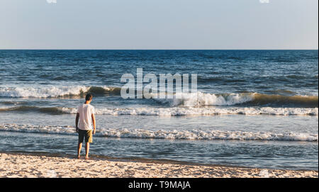 Junger Erwachsener Mann stand auf Sand und starrte auf kleinen Wellen des Meeres. Friedliche Atmosphäre im Sommer am Nachmittag. Ansicht von hinten. Leeren Platz für Text, Angebot oder Stockfoto
