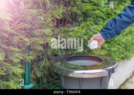 Ein jugendlicher Freiwilliger in einer blauen Jacke windbreaker wirft Kunststoff Papierkorb in einem Container vor dem Hintergrund eines grünen Zaun von thuja gesammelt. Die Stockfoto