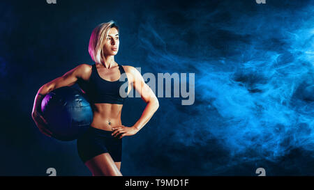 Sportliche Blondine in modischer Sportswear mit Medizinball posieren. Foto von muskulöse Frau auf dunklem Hintergrund mit Rauch. Kraft und Motivation. Stockfoto