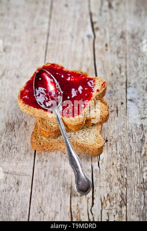 Geröstetes Getreide Scheiben Brot mit selbstgemachter Kirschmarmelade und Löffel closeup auf rustikalen Holztisch Hintergrund stack. Süße Speisen zum Frühstück. Stockfoto
