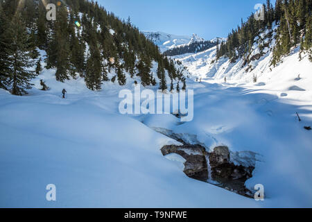 Skifahrer wandern die Asulkan Tal auf den Gletscher in der Ferne. Ein Wasserfall auf seine Rechte aus dem Frühling Schnee geschmolzen ist. Stockfoto