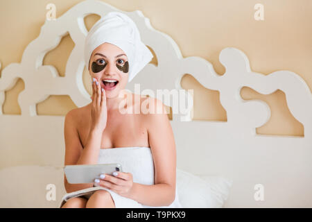 Schön überrascht oder junge Frau mit unter Auge Patches schockiert und Reden Handy in Bademantel im Bett lag. Happy girl kümmert sich Stockfoto