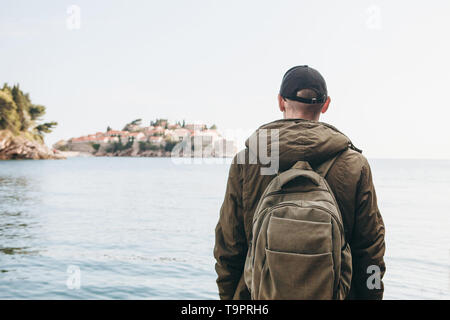 Touristen mit einem Rucksack in der Nähe des Meeres. Allein reisen. In die Ferne schaut. Vor uns liegt die Insel Sveti Stefan in Montenegro. Stockfoto