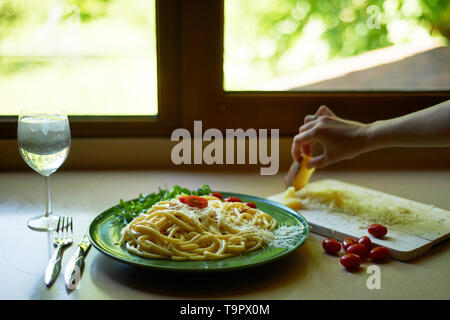 Pasta Carbonara mit geriebenem Parmesan und Cherry Tomaten, dekoriert mit Rucola. italienischen Mittagessen. Die Hand einer Frau setzt ein Stück Käse. Stockfoto