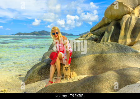 Lächelnd lifestyle Frau sitzt auf einem Felsbrocken, berührt einen niedlichen Hund an der Anse Source D'Argent einem der schönsten Strände von La Digue, Seychellen Stockfoto