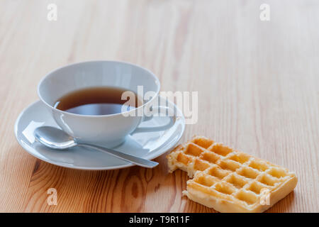 Hohe Betrachtungswinkel und einer weißen Teetasse, Untertasse und Löffel, sowie der angrenzenden gebissen Waffel und Krümel auf Weichholz Tisch. Stockfoto