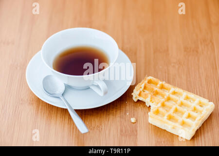 Hohe Betrachtungswinkel und einer weißen Teetasse, Untertasse und Löffel, sowie der angrenzenden gebissen Waffel und Krümel auf Weichholz Tisch. Stockfoto