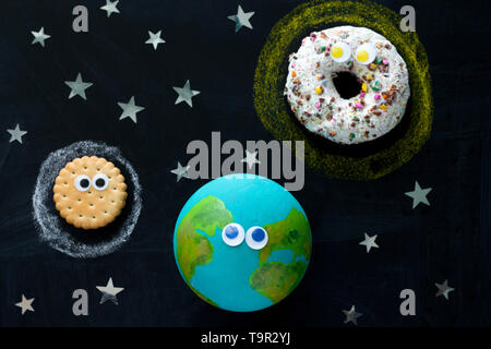 Cute Modell der Erde, Sonne und Mond mit kulleraugen auf dem Schwarzen Brett ein Donut, eine Kugel und einen Cookie auf dem Schwarzen Brett Stockfoto
