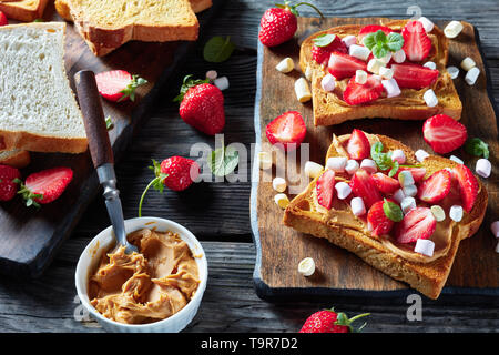 Sandwiches von geröstetem Brot mit Erdnussbutter, in Scheiben geschnittene Erdbeeren und Marshmallows auf einem hölzernen Schneidebrett auf einem alten rustikalen Tisch, cllose-up Stockfoto